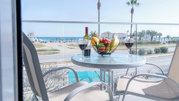 Un balcon avec une table dressée pour deux personnes, agrémentée d’un panier de fruits, de deux verres à vin et d’une bouteille de vin. La vue comprend une plage et des palmiers.