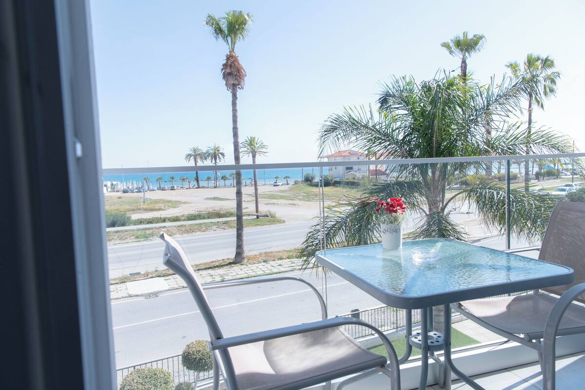 Un balcón con una mesa y sillas de vidrio, con vista a una calle con palmeras y una vista de la playa.