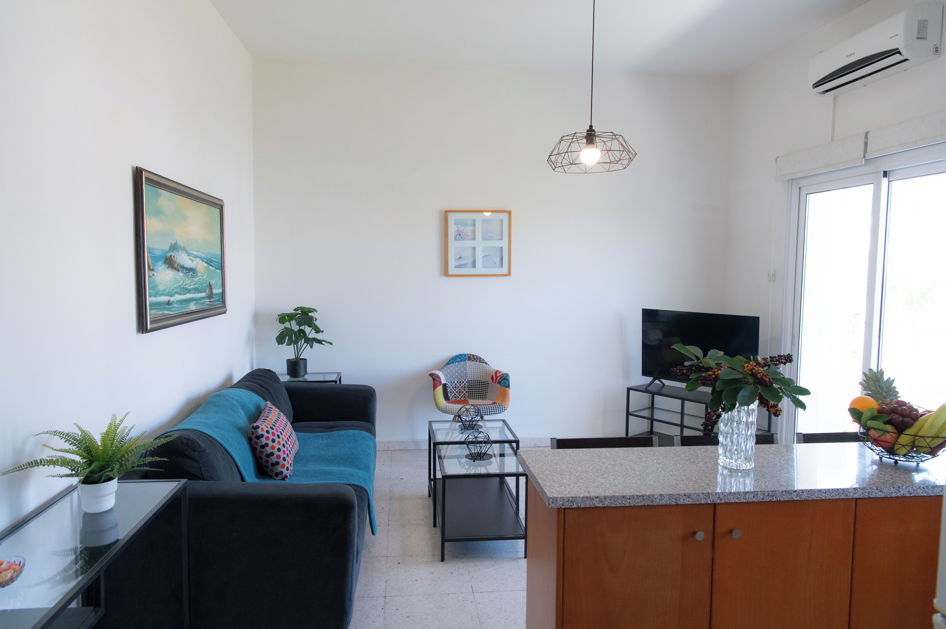 Una sala de estar moderna con un sofá, una mesa de centro de vidrio, un televisor y una zona de cocina con un frutero.