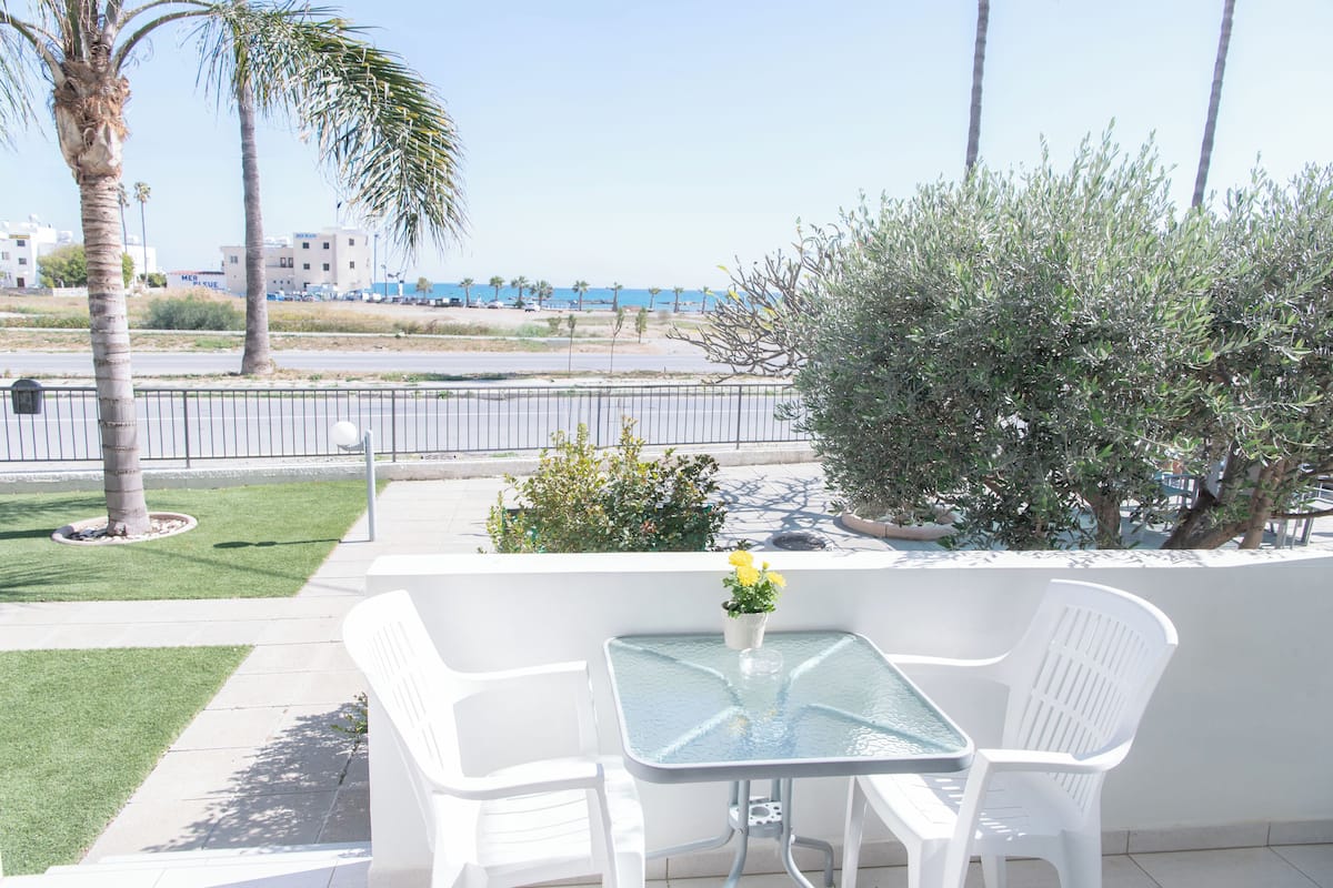 Un balcón con una mesa de vidrio y sillas blancas, con vista a una playa y palmeras.