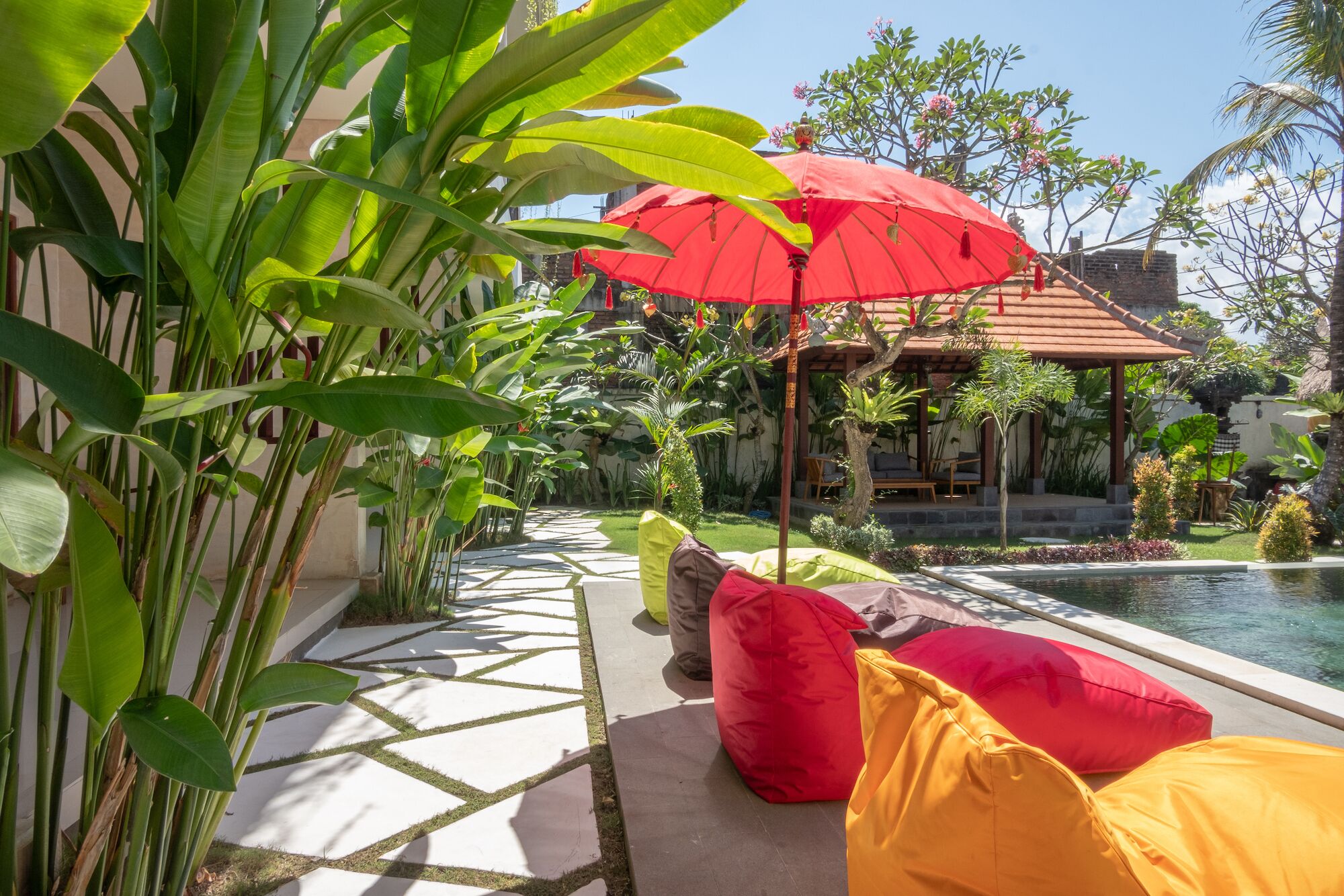 A poolside area with a geometric stone pathway, a covered seating area with red and yellow cushions, and lush tropical plants.