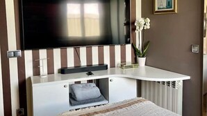 Ein Hotelzimmer mit einem großen Flachbildfernseher an einer Wand mit gestreifter Tapete, einem weißen Konsolentisch mit Flachbildfernseher, einem Bett mit einer beigefarbenen Decke und einer Topfpflanze.