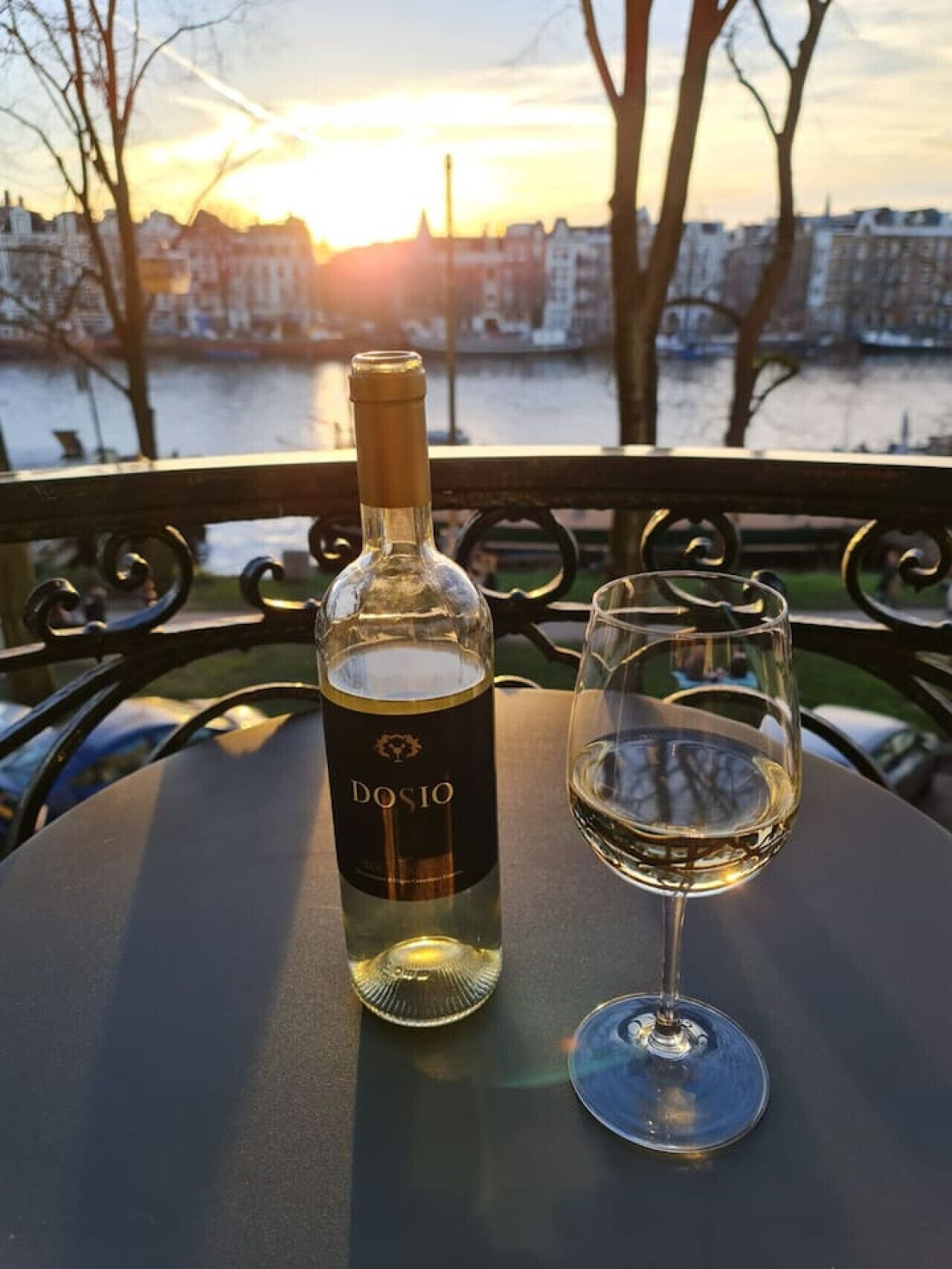 A bottle of Dosio wine and an empty glass on a table with a view of a river and buildings in the background.