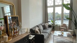 A modern living room with a grey sofa, a coffee table, a black cabinet with wine bottles, a large mirror, and a view of a cityscape through large windows.