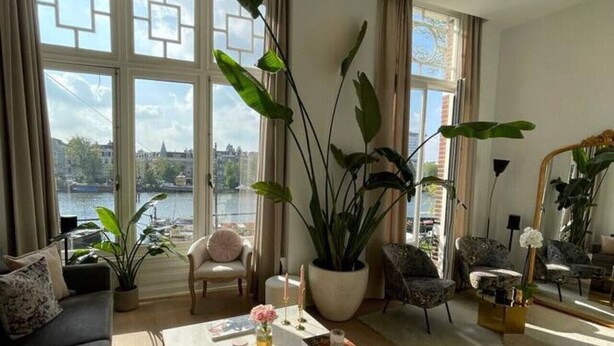 A spacious living room with a large window overlooking a river, a sofa, a coffee table with magazines, and several potted plants.