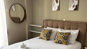 Una cama bien hecha con sábanas blancas y dos almohadones decorativos, un espejo redondo en la pared y una lámpara en el techo.