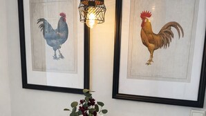 A round wooden table with a black vase, two framed rooster illustrations, and a white device on the wall. - Classic Comfort in the Heart of Monastiraki | Walk to the Acropolis, Agora & More (Athina)