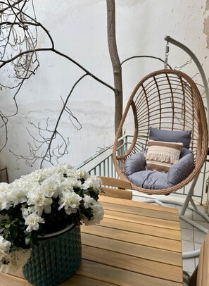A wicker hanging chair with cushions, a wooden table, and a potted plant with white flowers. - Elegant Kolonaki Retreat | Sophisticated 2-Bedroom Apartment in Upscale Central Athens (Athina)