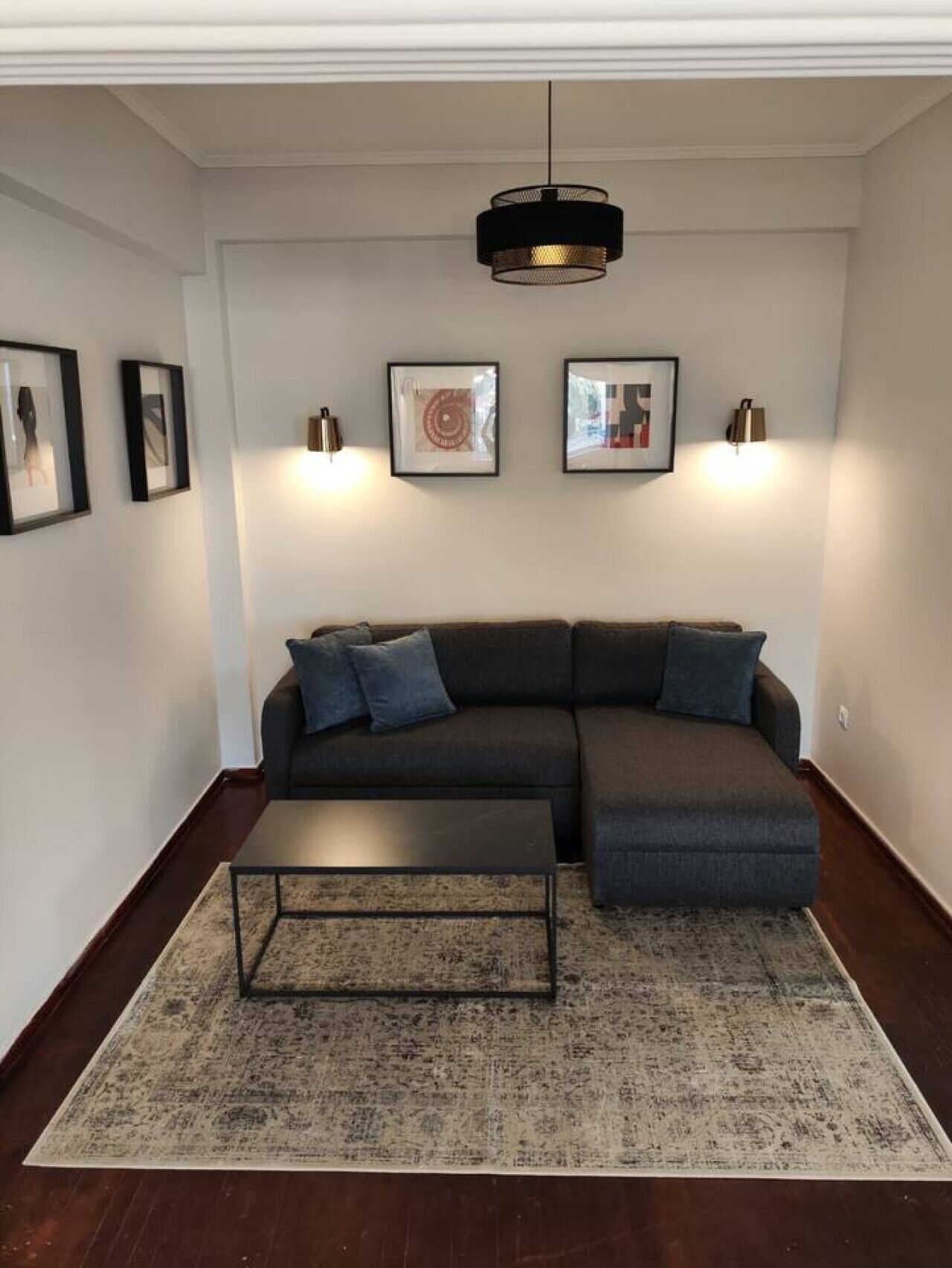A modern living room with a dark gray sectional sofa, a small coffee table, and a patterned rug. There are three framed artworks on the wall, and a pendant light hangs above the sofa.