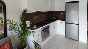A modern kitchen with white cabinets, a stainless steel refrigerator, and a built-in oven. There is a grey sofa with a red cushion and a potted plant in the corner. - Sunset Sea View Pangrati Flat (Athina)