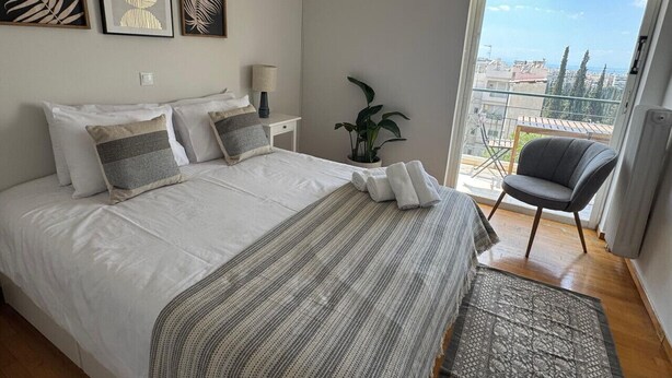A bedroom with a large bed, a small table with a lamp, a chair, and a view of the city through a balcony. - Sunset Sea View Pangrati Flat (Athina)