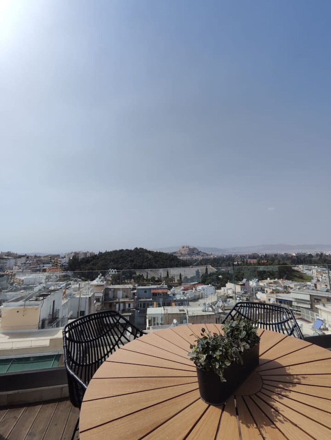 A rooftop terrace with a wooden table and chairs, a potted plant, and a view of a cityscape with buildings and a hill in the distance.