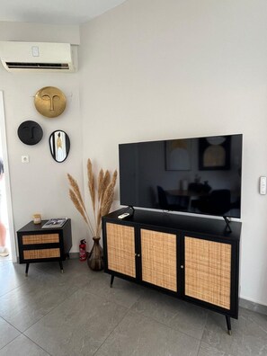 A living room with a flat-screen TV, a black cabinet with wicker doors, a small side table, and decorative wall art. - Rooftop Serenity in Pangrati | Jacuzzi, 2 Terraces & Acropolis Views in Central Athens (Athina)
