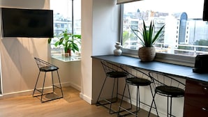 A modern kitchen with a large window, bar stools, a flat-screen TV, and a potted plant.