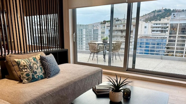 A modern living room with a sofa, a coffee table, and a view of a cityscape through large windows.