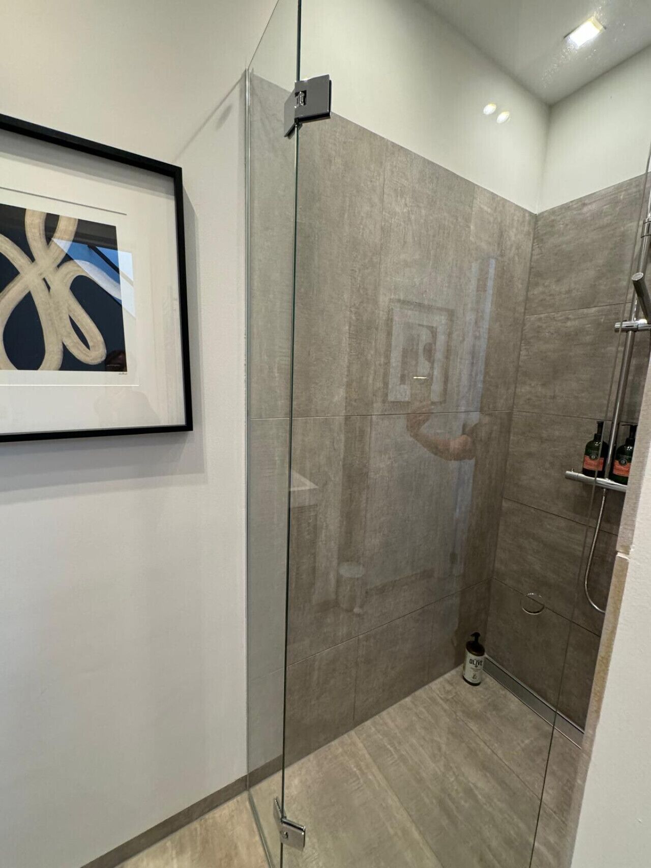 Une salle de bain moderne avec une douche à parois de verre, une œuvre d’art abstraite encadrée accrochée au mur et une petite bouteille d’huile d’olive posée sur le sol.