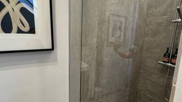 Une salle de bain moderne avec une douche à parois de verre, une œuvre d’art abstraite encadrée accrochée au mur et une petite bouteille d’huile d’olive posée sur le sol.