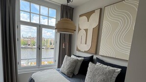 A modern bedroom with a large bed, a view of a cityscape through a window, and abstract wall art. - Grand Amstel Residence | Expansive Two-Story Riverfront Apartment in Central Amsterdam (Amsterdam)