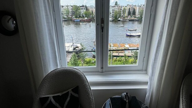 A room with a curved window overlooking a river, a chair, a small table, and a clock on the wall. - Grand Amstel Residence | Expansive Two-Story Riverfront Apartment in Central Amsterdam (Amsterdam)
