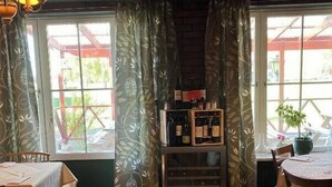 A cozy dining area with wooden tables and chairs, large windows with patterned curtains, and a wine rack against the wall. - Brevensgården (Brevens Bruk)