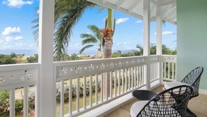 A covered outdoor area with white railings, two black wire chairs, and a view of palm trees and a garden. - Greens 2BR (Montego Bay)