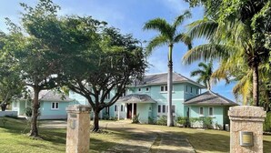 A light blue two-story house with a front yard, palm trees, and a stone pillar with a plaque. - Greens 2BR (Montego Bay)