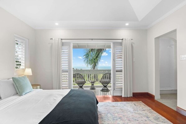 A bedroom with a bed, a nightstand, a lamp, a balcony with chairs, and a view of the ocean.