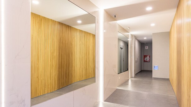 A modern hallway with a wooden slatted wall, a glass elevator, and a fire extinguisher on the wall.