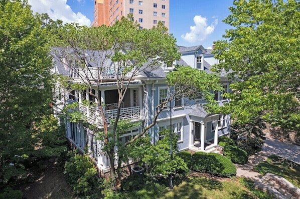 A two-story house with a balcony, surrounded by lush greenery and a clear sky.