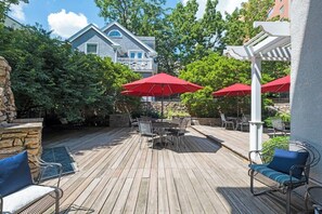 A wooden deck with outdoor furniture and red umbrellas, surrounded by trees and a stone wall.