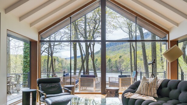 Ein geräumiger Raum mit großen Fenstern und Blick auf bewaldetes Gelände sowie Berge. Es gibt einen Glastisch, ein dunkles Sofa und eine Holzbank.
