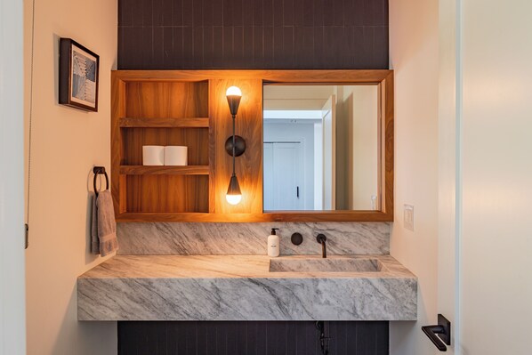 Un baño con encimera de mármol, estantes de madera, un espejo grande y una lámpara de pared.