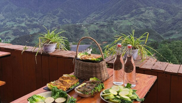 Mesa de madera dispuesta con comida y bebidas, rodeada de plantas en macetas, con vistas a un paisaje montañoso.