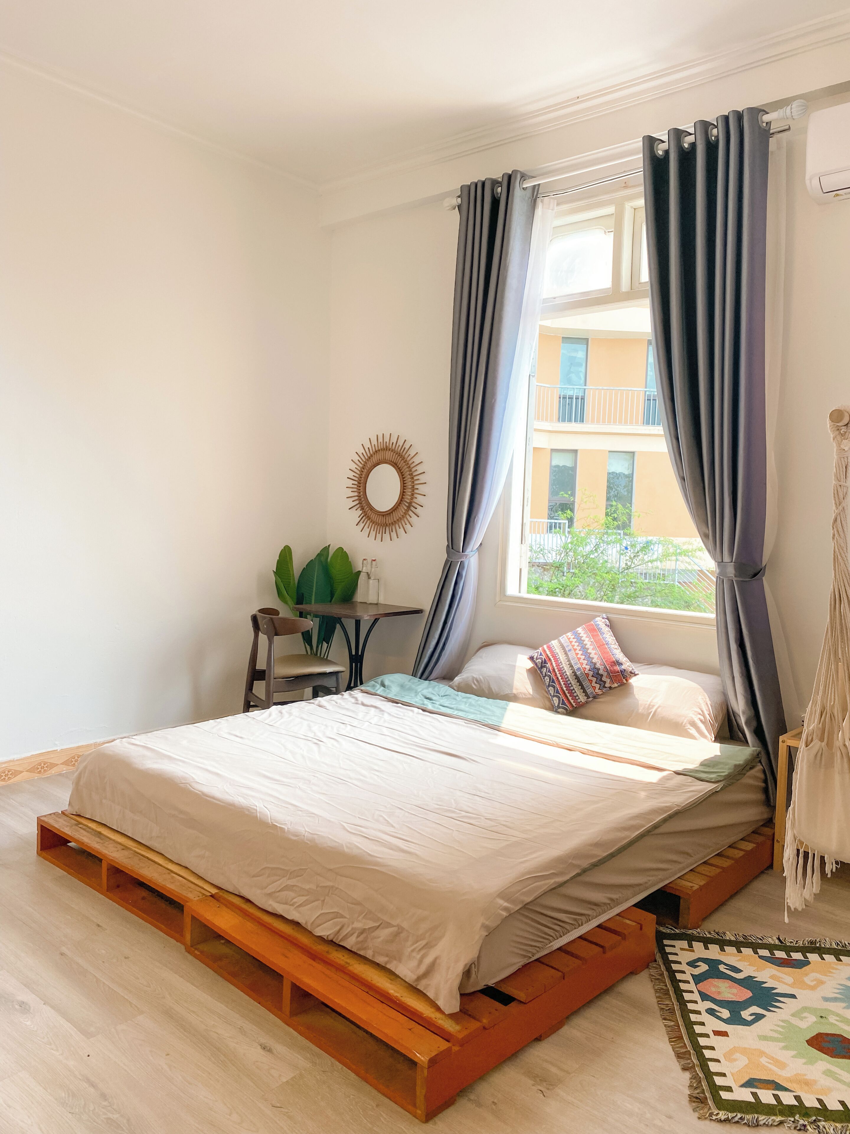 Ein Schlafzimmer mit hölzernem Bettgestell, einem kleinen Tisch mit Pflanze und Blick auf ein Gebäude.