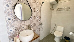 Un baño con un espejo redondo, un lavabo con estante de madera, un inodoro y una ducha con cabezal de ducha manual.