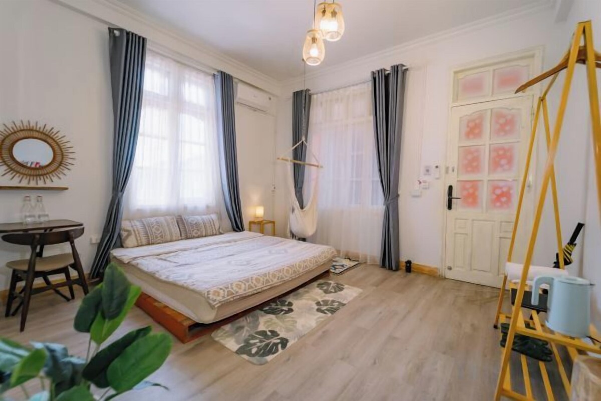 A bedroom with a bed, a desk, and a chair. There is a window with curtains, a door with a window, and a wall-mounted mirror.