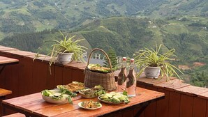 Una mesa de madera con diversos alimentos y plantas en macetas en un balcón con vistas a un paisaje montañoso.