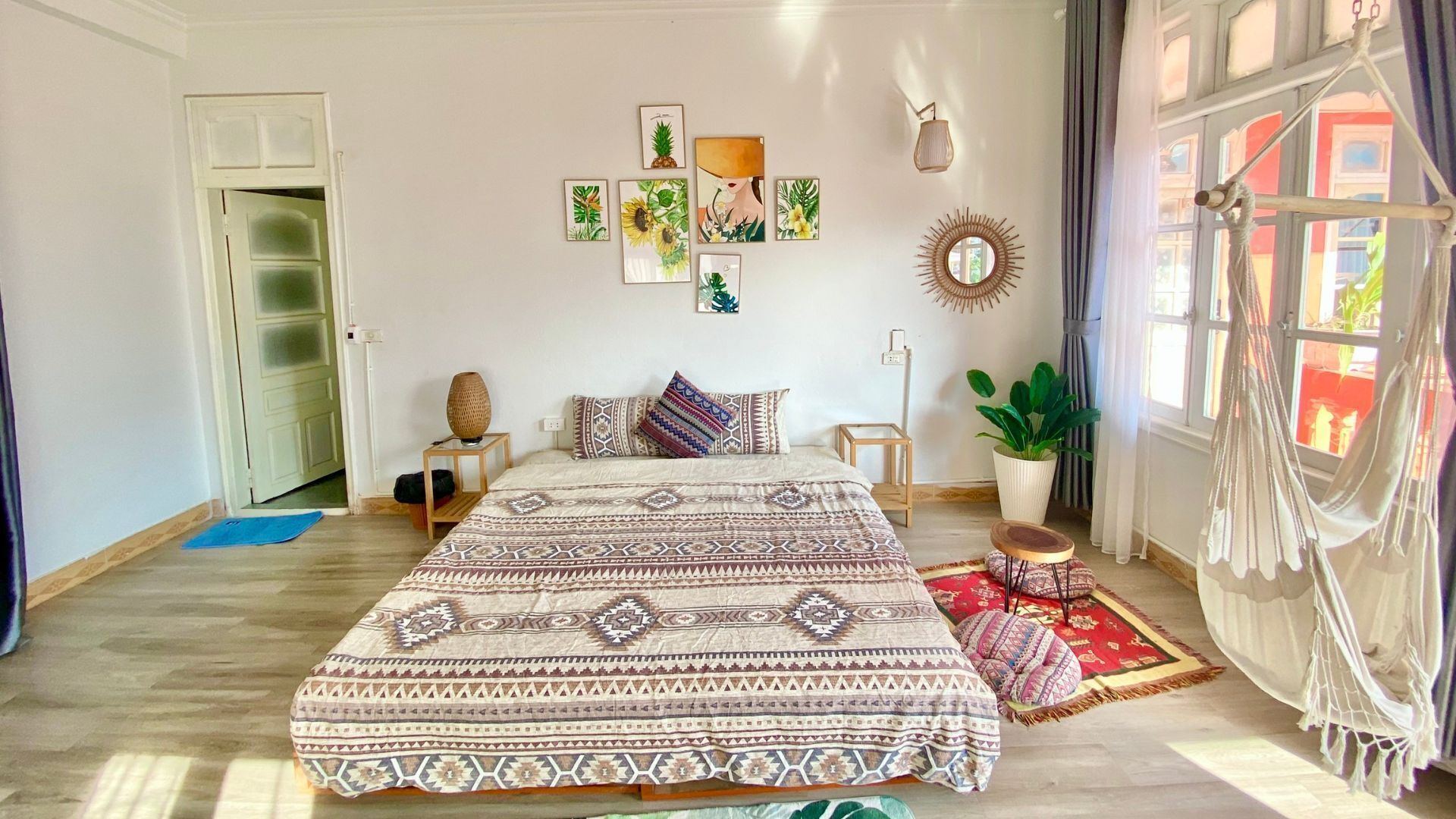 A spacious bedroom with a large bed, a wooden nightstand, a potted plant, and a view of the outdoors through a window with curtains.