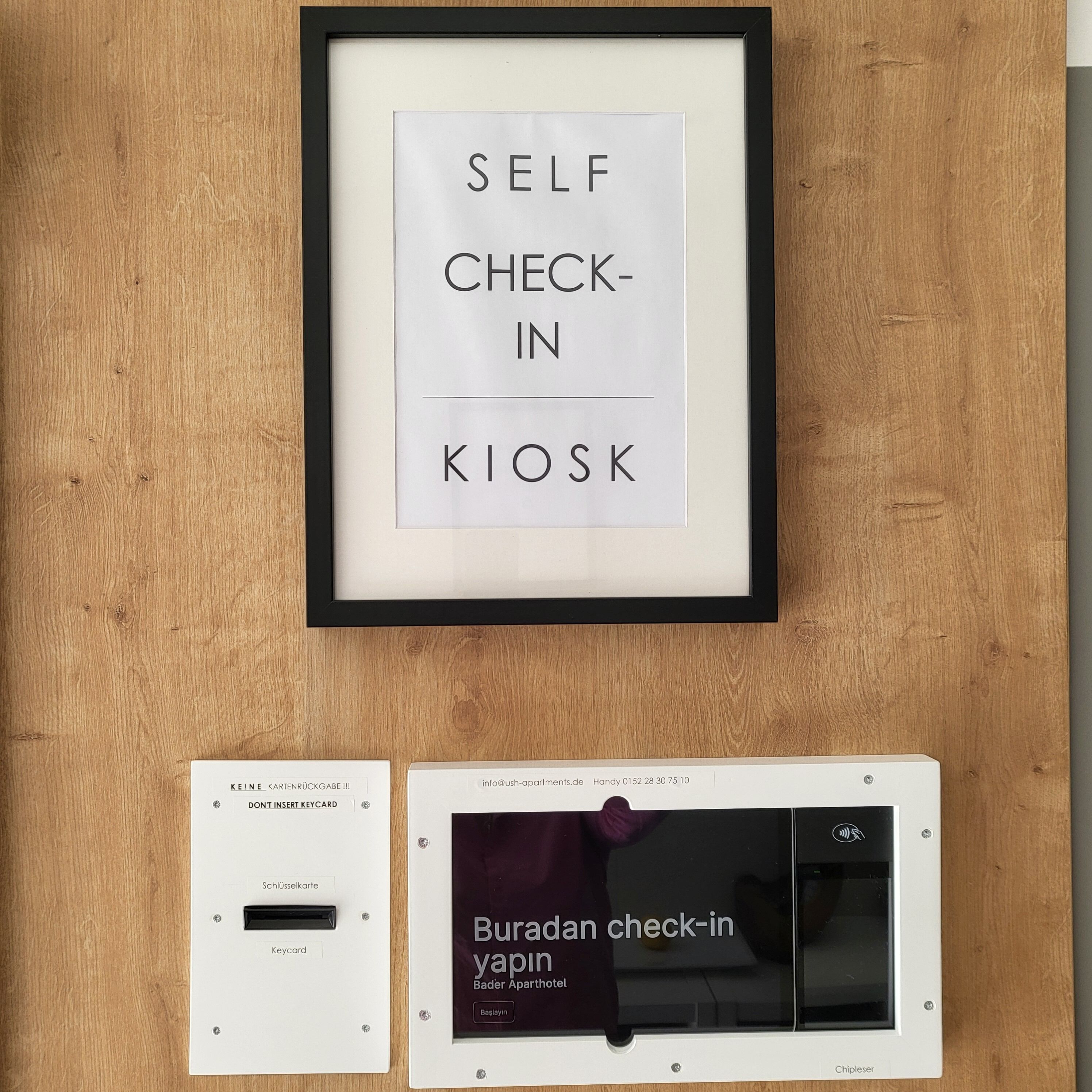 Una postazione self-check-in con un cartello incorniciato sopra e un lettore di tessere chiave sottostante, e un display digitale per il check-in della stanza.