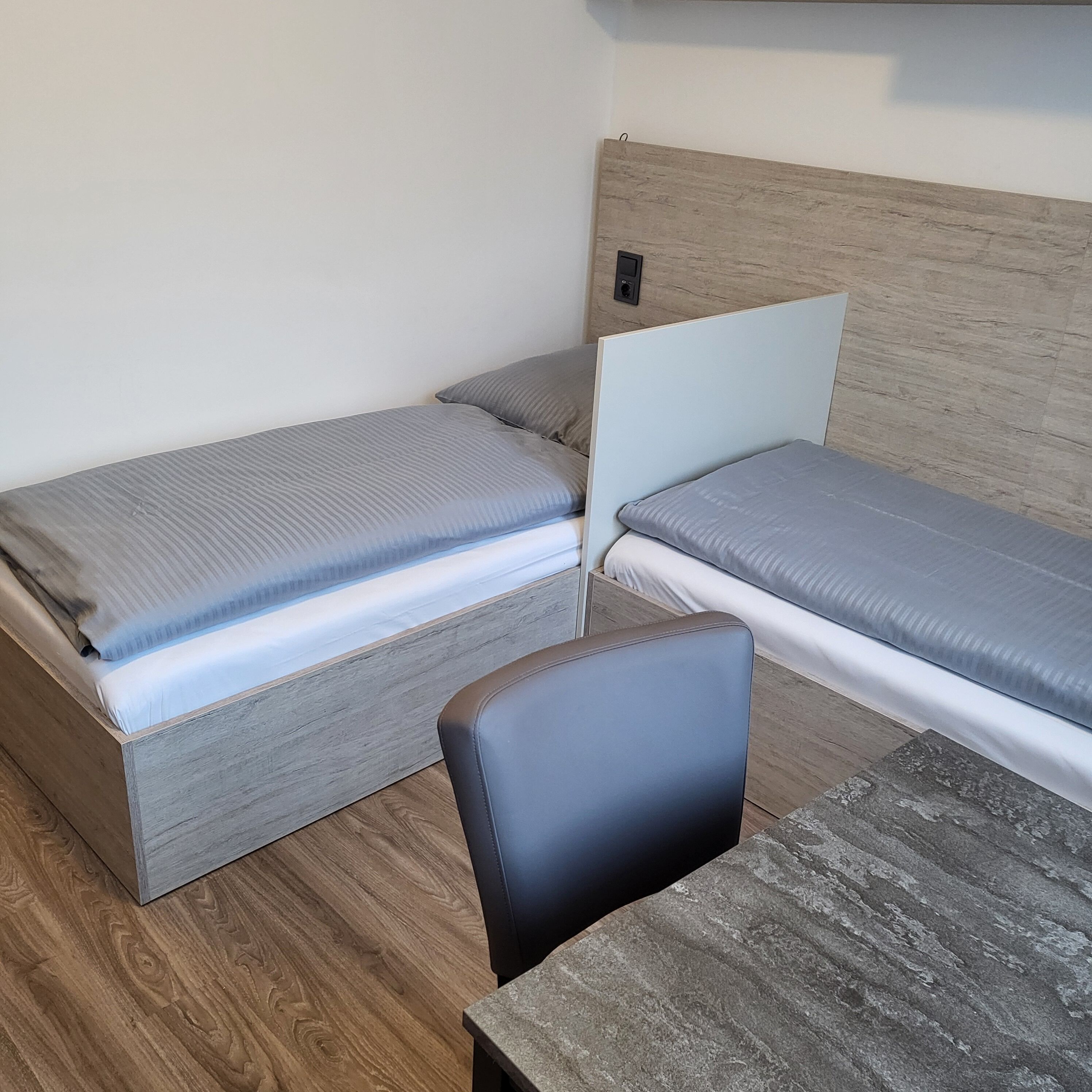 Una habitación pequeña y funcional con dos camas individuales, un cabecero de madera, una mesita y una silla.