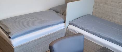 Una habitación pequeña y funcional con dos camas individuales, un cabecero de madera, una mesita y una silla.