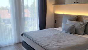 A modern bedroom with a large bed, a wooden headboard, and a view of the outside through a window with sheer curtains. - Bader Aparthotel (Unterschleißheim)