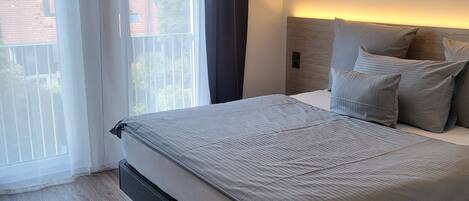 A modern bedroom with a large bed, a wooden headboard, and a view of the outside through a window with sheer curtains.