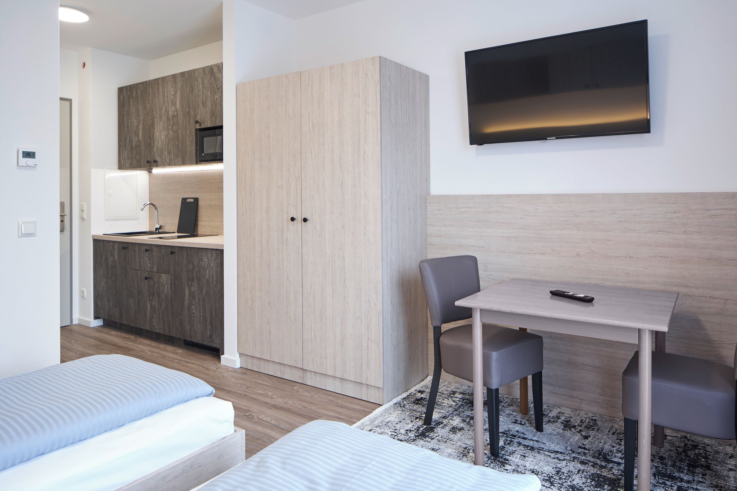 Une chambre d’hôtel moderne équipée d’une kitchenette, d’un téléviseur fixé au mur, d’un lit, d’un bureau avec une chaise et d’une petite table.