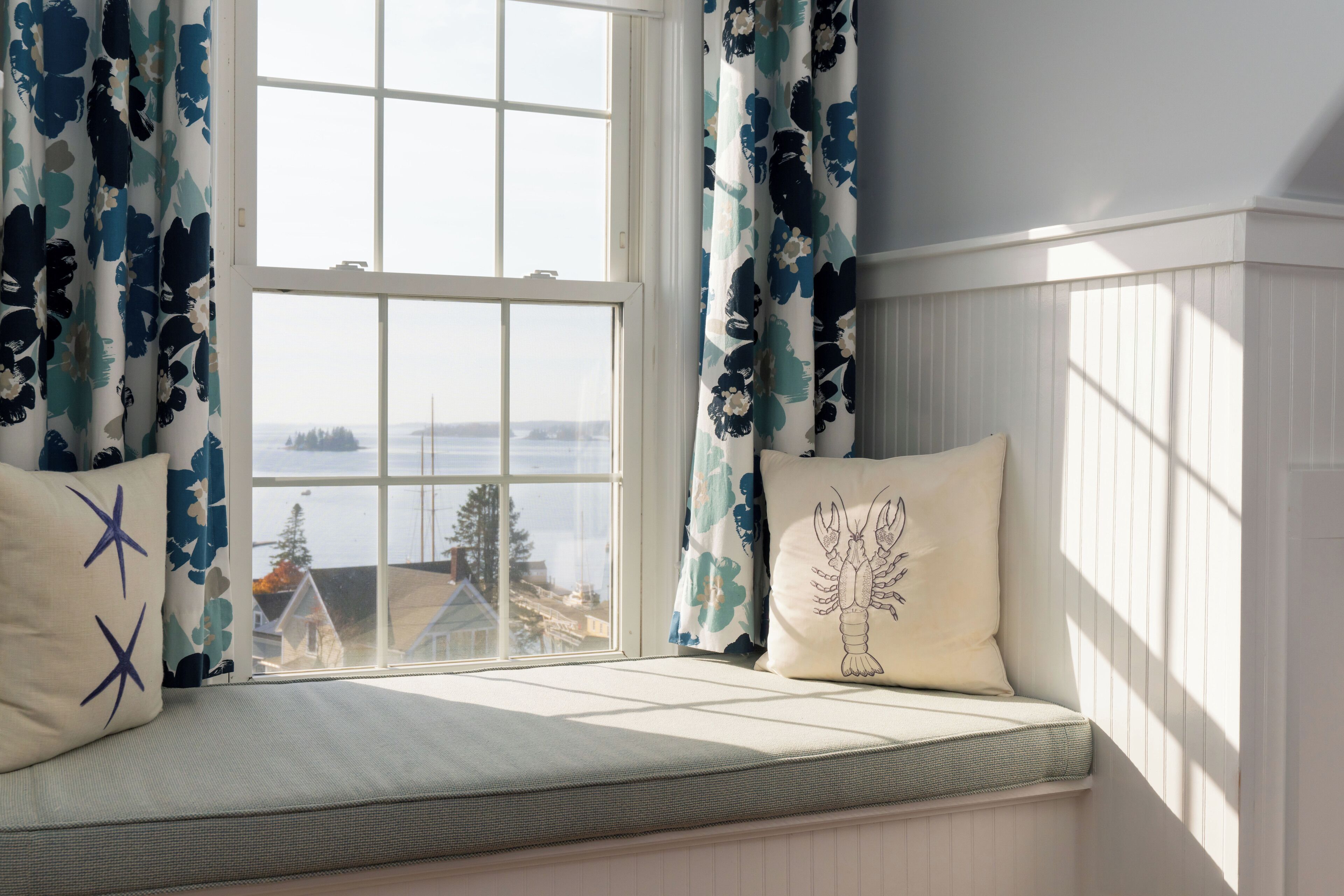 A bay window with a view of a house and trees, a cushioned bench, and patterned curtains.