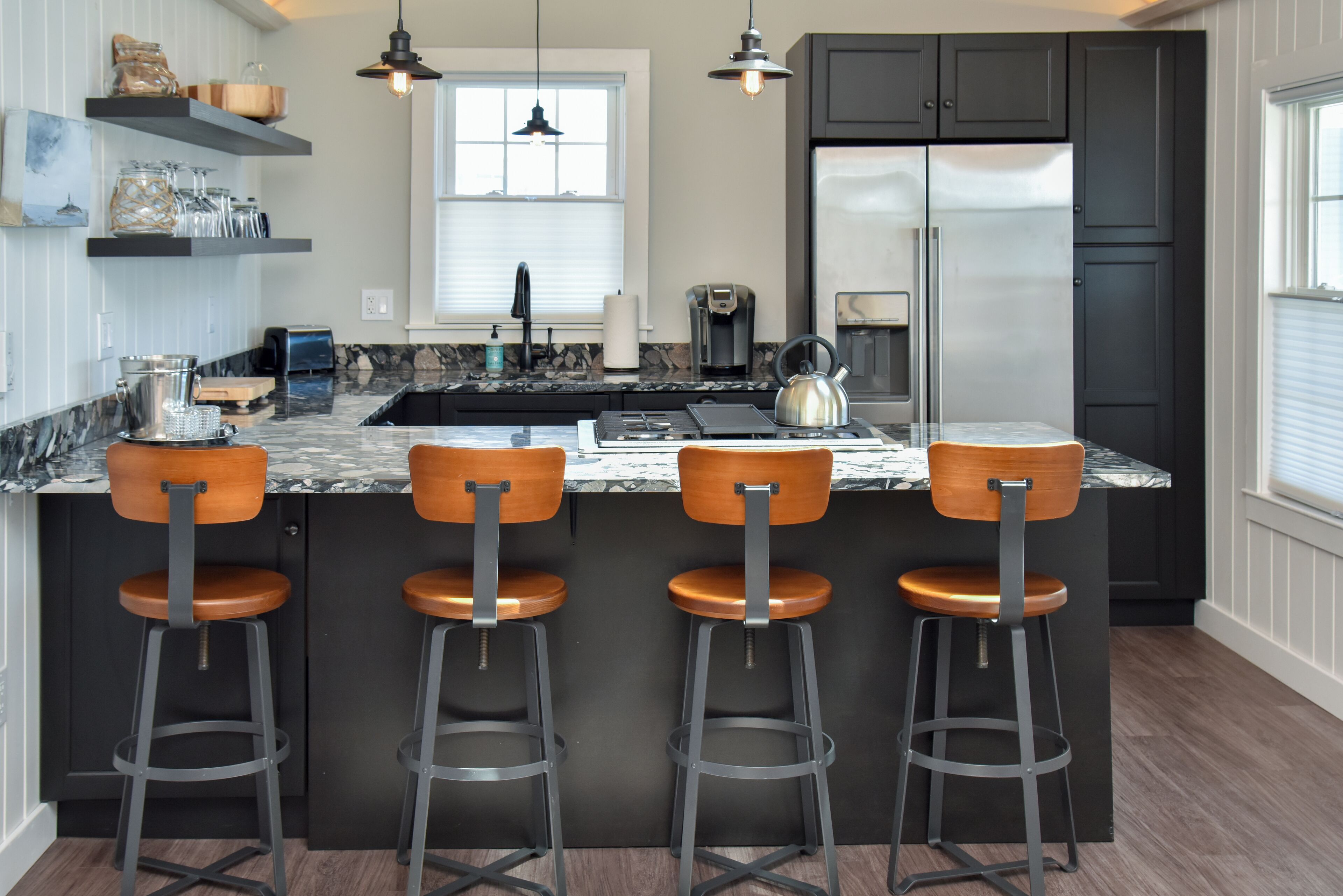 A modern kitchen with a central island, bar stools, a refrigerator, and a coffee maker.