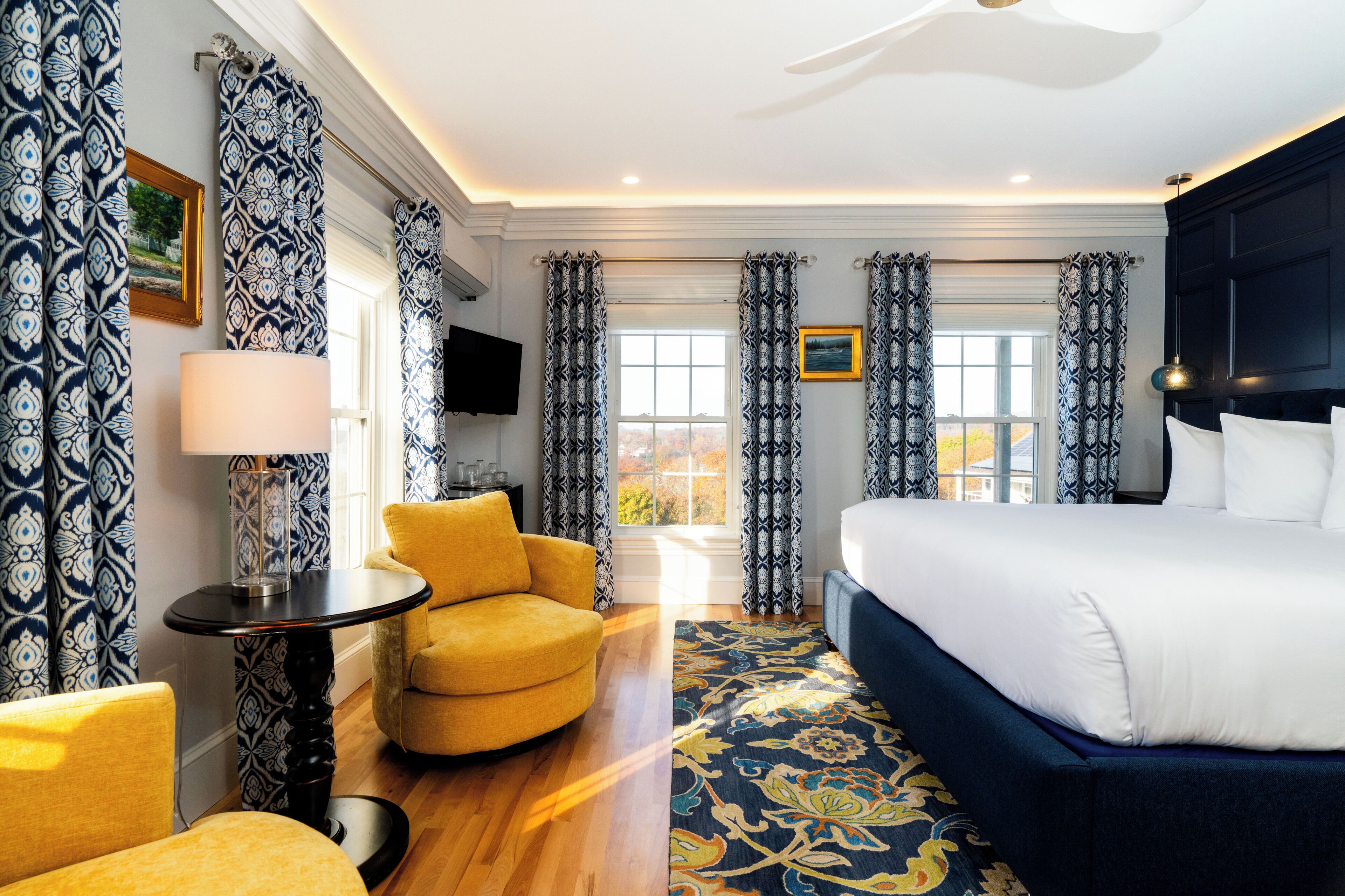 A hotel room with a large bed, a round table with a lamp, a yellow armchair, and patterned curtains.