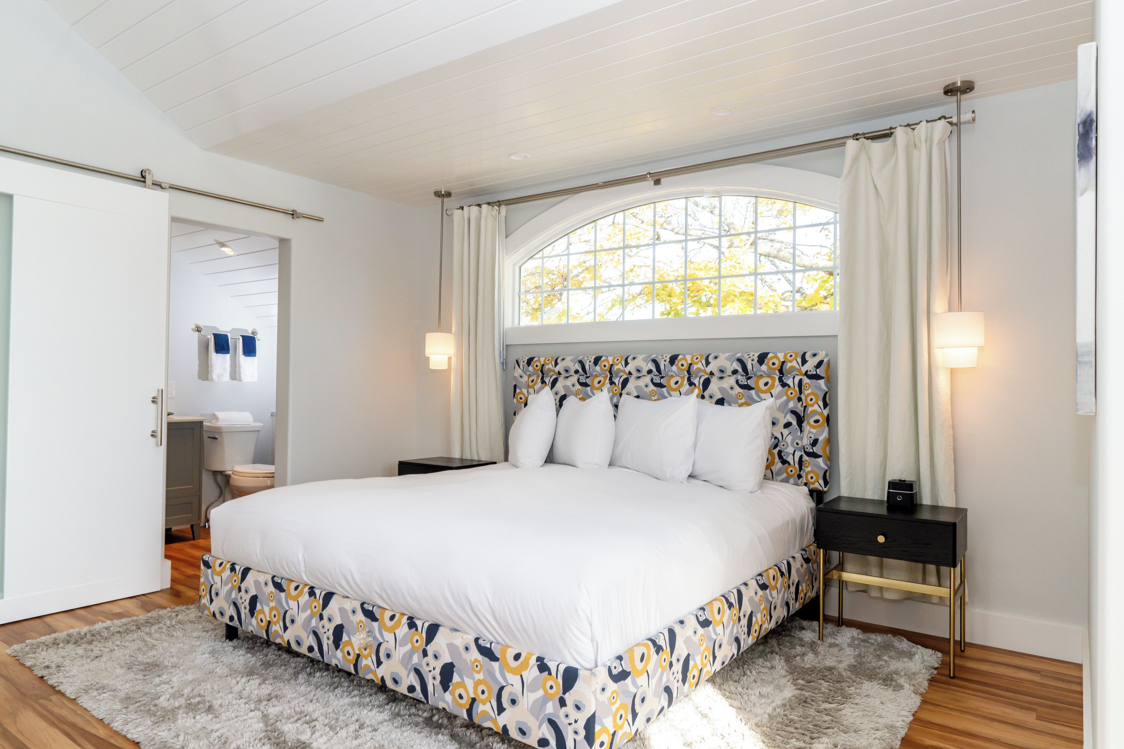 A bedroom with a large bed, a nightstand, a window with curtains, and a door leading to a bathroom.