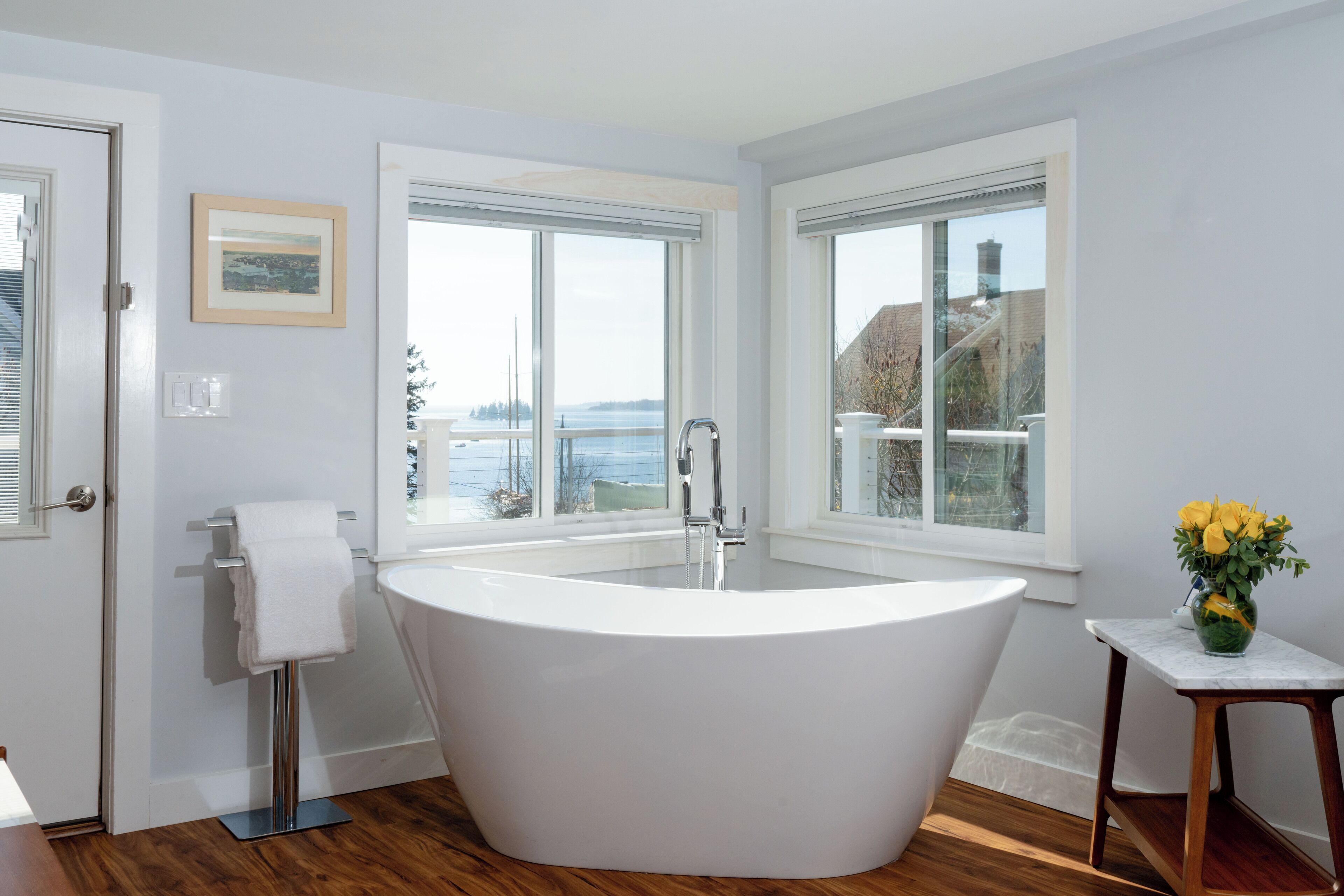 Un baño moderno con una bañera independiente, ventanas amplias que ofrecen vista al mar y una pequeña mesa con un jarrón de flores.