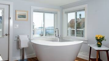 Un baño moderno con una bañera independiente, ventanas amplias que ofrecen vista al mar y una pequeña mesa con un jarrón de flores.